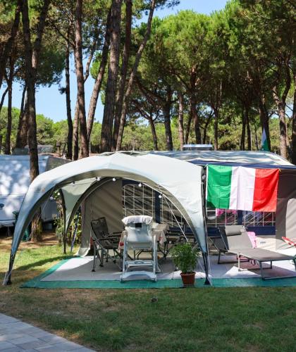 Camping mit italienischer Flagge, Stühlen und Liegen unter Bäumen.