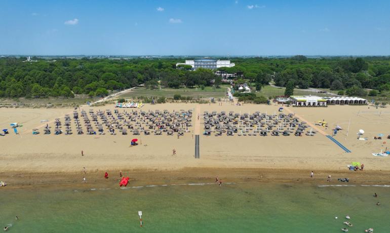 Spiaggia affollata con ombrelloni, mare calmo e vegetazione sullo sfondo.