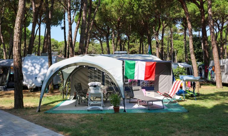 Campeggio con bandiera italiana, sedie e lettini sotto gli alberi.