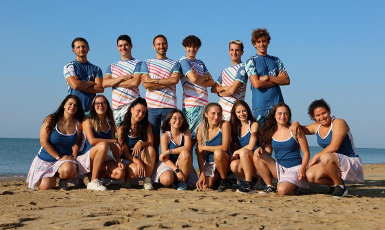 Gruppe på stranden i farverigt tøj, smilende under klar himmel.