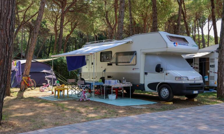 Campingvogn på campingplads med cykler og bord under træer.